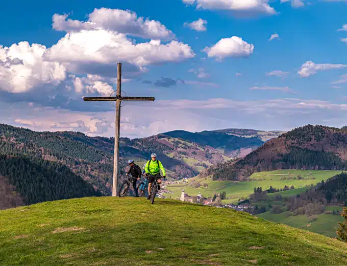 Radtour im Münstertal – Mountainbiker an einem Gipfelkreuz und Blick über das grüne Tal
